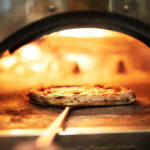 This photo shows a pizza with a beautifully blistered crust being peeled out of a wood-fired oven.