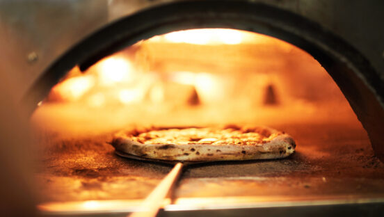This photo shows a pizza with a beautifully blistered crust being peeled out of a wood-fired oven.