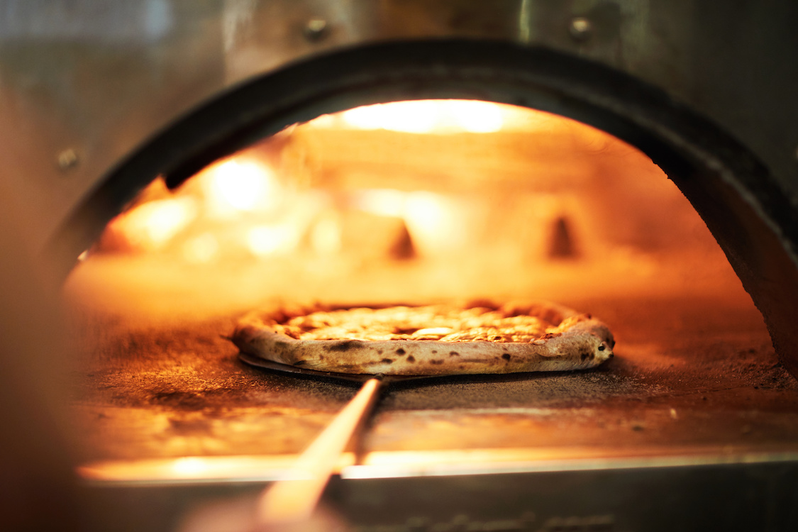 This photo shows a pizza with a beautifully blistered crust being peeled out of a wood-fired oven.
