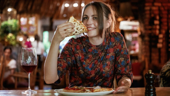 This photo shows a young woman smiling and eating a slice of pizza in a pizzeria, with a plate containing the rest of the pizza on the table in front of her.