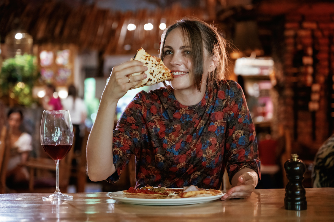 This photo shows a young woman smiling and eating a slice of pizza in a pizzeria, with a plate containing the rest of the pizza on the table in front of her.