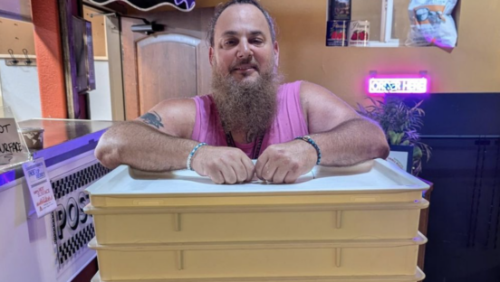 A man stands, smiling at the camera, with his arms on top of a series of tubs that hold pizza dough.