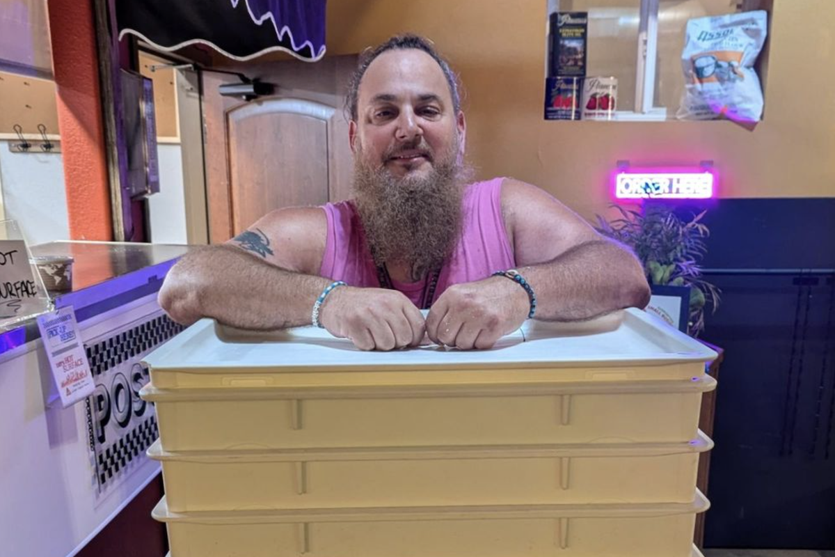 A man stands, smiling at the camera, with his arms on top of a series of tubs that hold pizza dough.