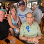 This photo shows Ron Help, wearing a light green shirt, glasses and a blue ribbon that reads, "Birthday Boy," at a birthday celebration with a group of friends or family members at his restaurant.