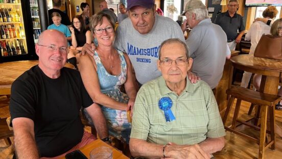 This photo shows Ron Help, wearing a light green shirt, glasses and a blue ribbon that reads, "Birthday Boy," at a birthday celebration with a group of friends or family members at his restaurant.