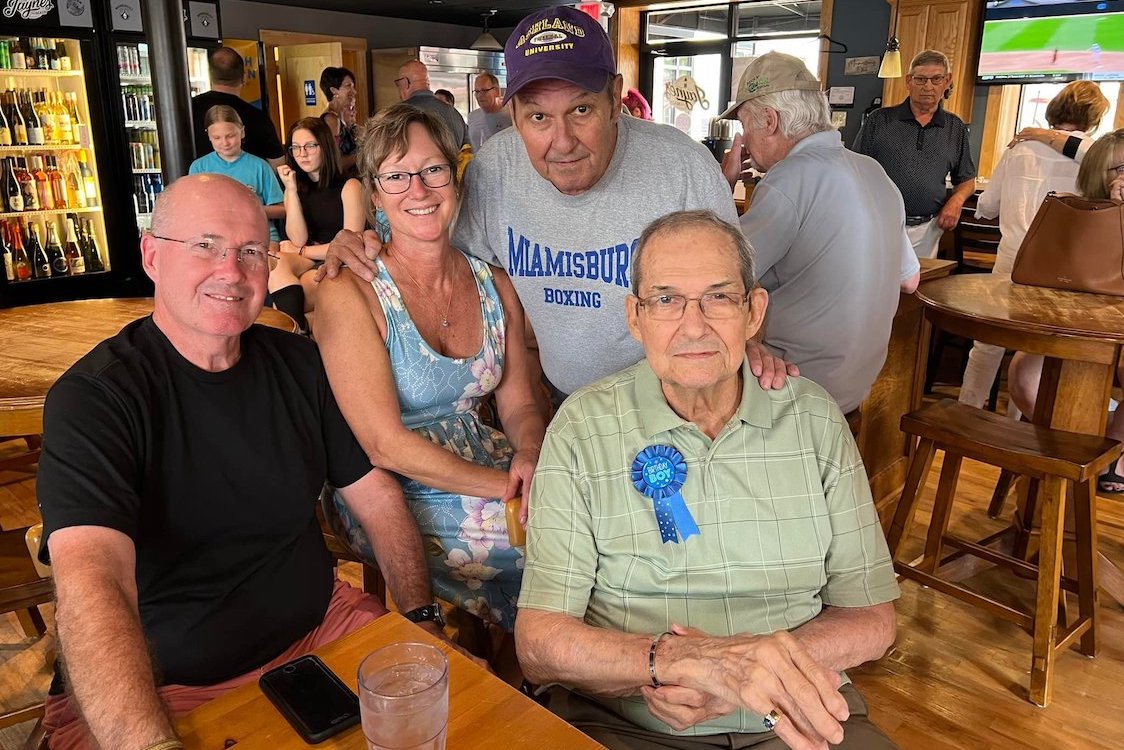 This photo shows Ron Help, wearing a light green shirt, glasses and a blue ribbon that reads, "Birthday Boy," at a birthday celebration with a group of friends or family members at his restaurant.
