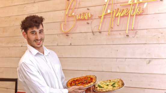 This photo shows a smiling, curly haired waiter holding two pizzas in a restaurant. Behind him is a neon sign that reads Buon Appetito!