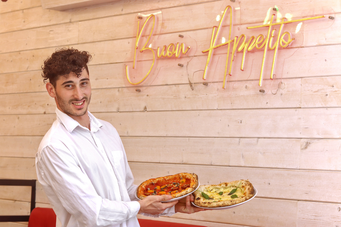 This photo shows a smiling, curly haired waiter holding two pizzas in a restaurant. Behind him is a neon sign that reads Buon Appetito!