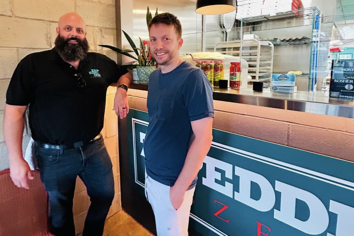 A middle-aged man and a young man stand in front of a pizza counter.