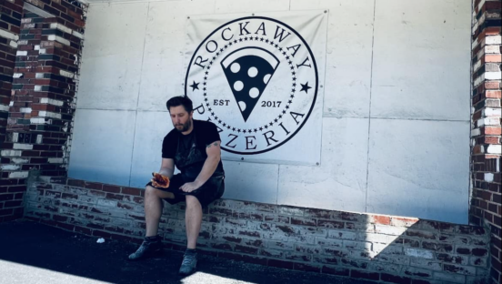 Josh Sickels, owner of Rockaway Pizzeria, sits in front of his shop in White Oak, PA, eating a piece of pizza.