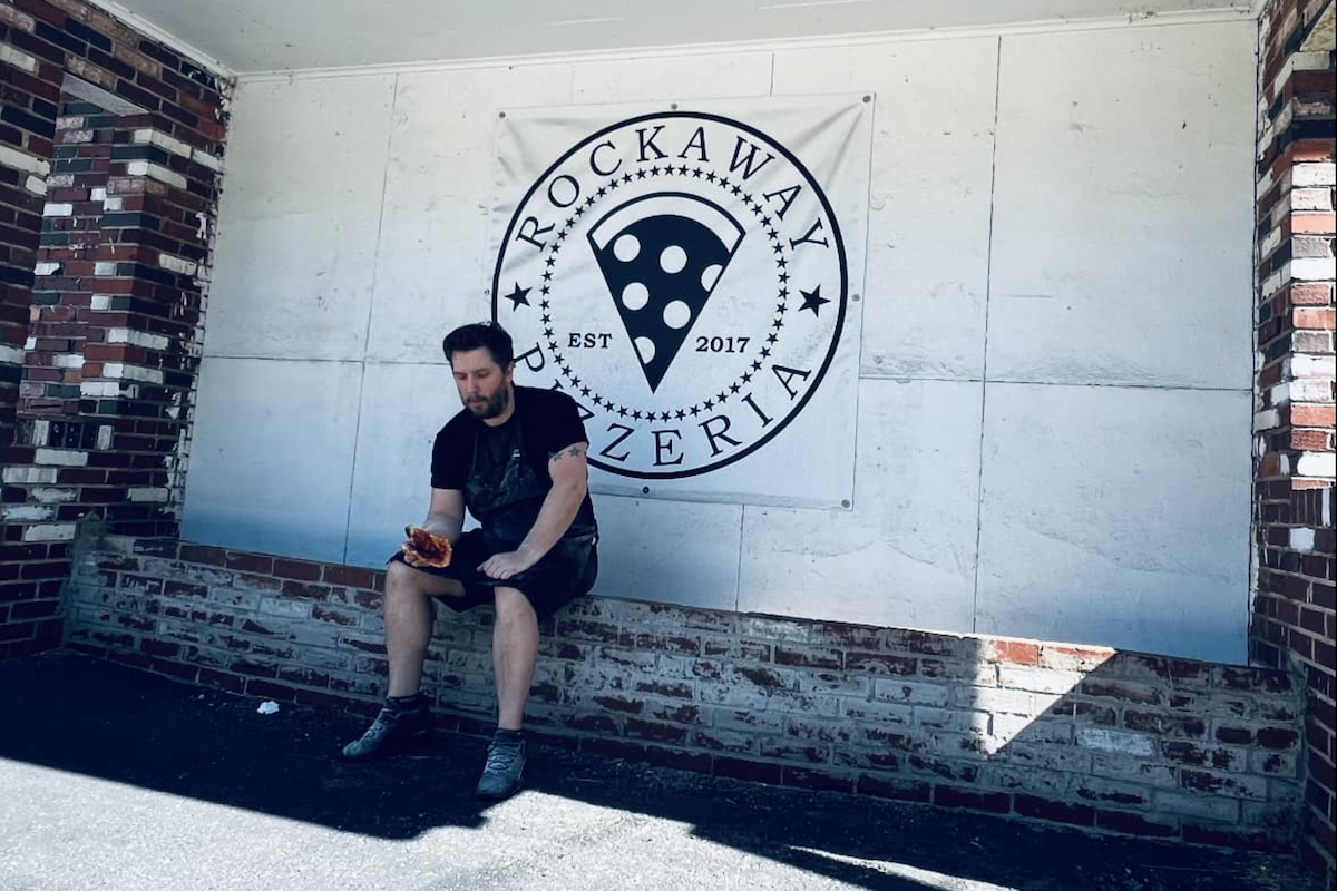 Josh Sickels, owner of Rockaway Pizzeria, sits in front of his shop in White Oak, PA, eating a piece of pizza.