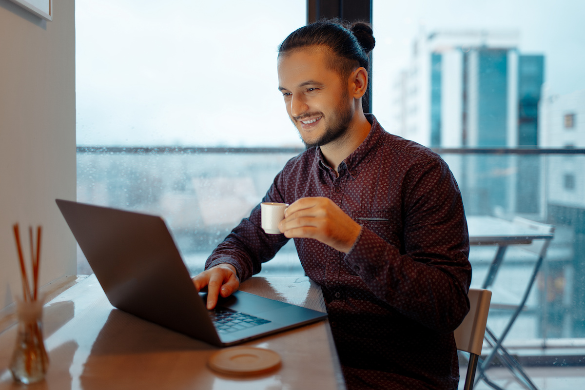 Portrait of handsome man working at laptop, typing on keyboard while holding a small cup of espresso coffee on background of panoramic windows.