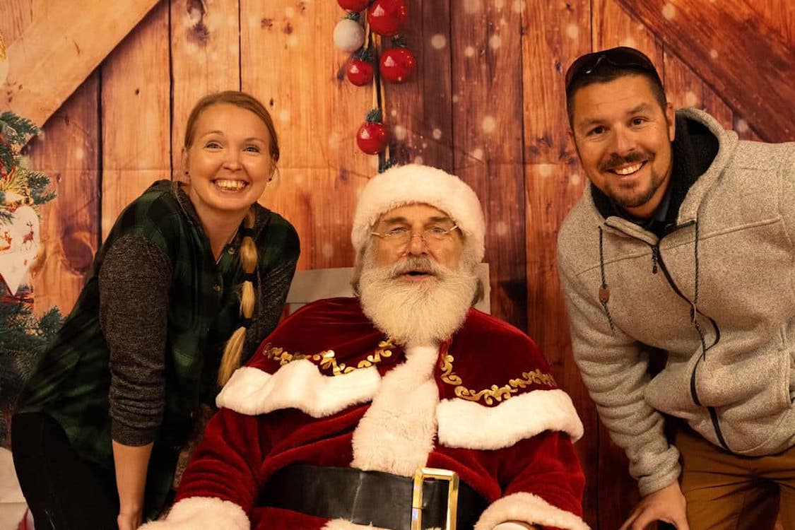 This warmly lit photo shows the owners of Cucinella's flanking Santa Claus, who is seated in front of a wood-paneled wall.
