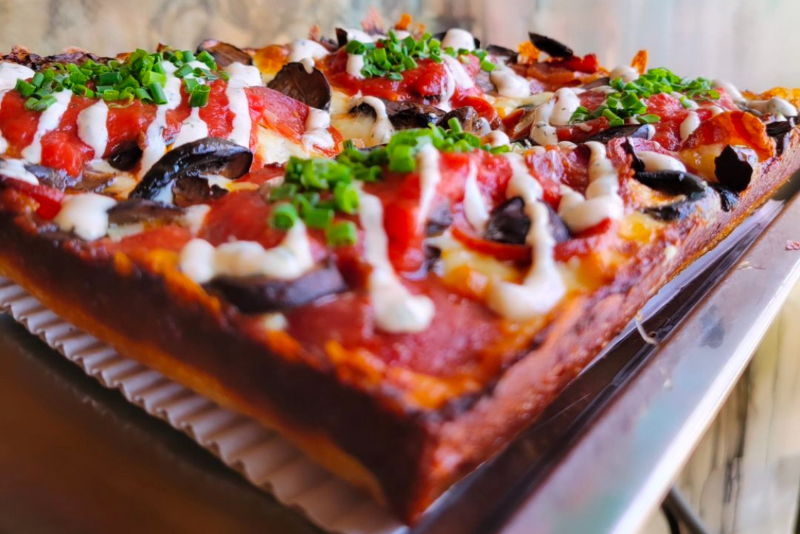 This photo shows a close-up of a Detroit-style pizza with pepperoni, mushrooms, chives and other ingredients.