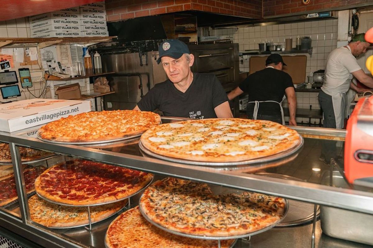 A pizza shop owner stands behind the counter with several full, New York-style pizzas in the foreground.