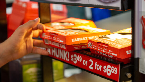 This photo shows a person's hand pulling a boxed Hunt Brothers pizza slice from a heating unit at a convenience store.