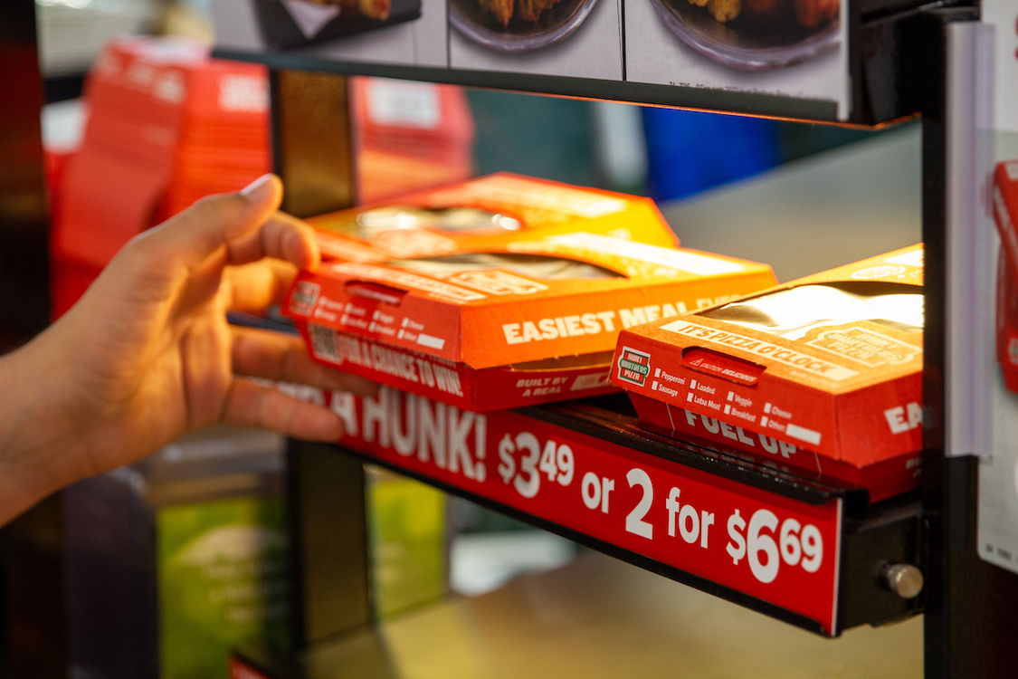 This photo shows a person's hand pulling a boxed Hunt Brothers pizza slice from a heating unit at a convenience store.