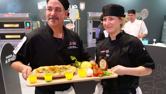 This photo shows Jamie Culliton and McKenna Carney, both dressed in black chef coats, showing a pizza at a culinary competition.