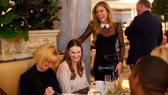 This photo shows a well-dressed Katie Lee standing over a table of customers at one of her ticketed events. She is laughing, and the customers look happy to be there.