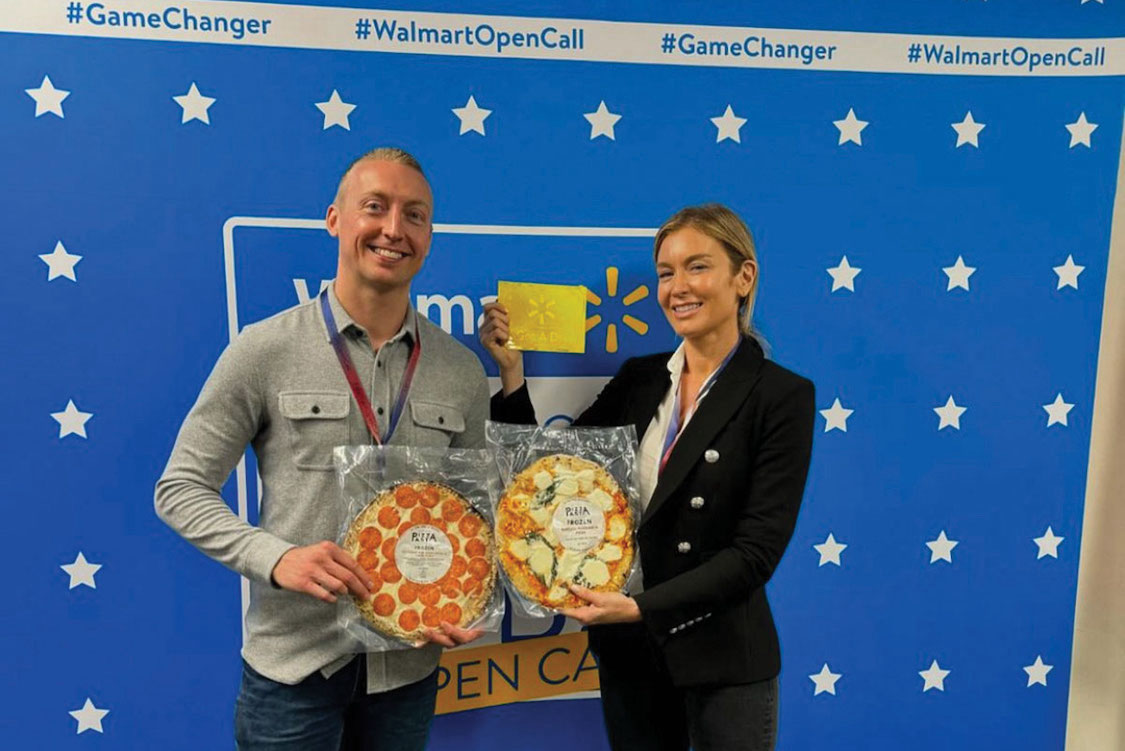 This photo shows Katie Lee on the right and Jake Sanderson on the left, holding up two of their frozen pizzas at the Walmart headquarters. They're standing in front of a blue banner with the words #GameChanger and #WalmartOpenCall.