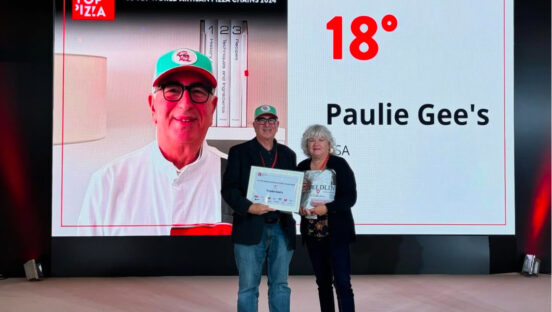 This photo shows Paulie Gee accepting his 50 Top Pizza award with a woman standing next to him. Behind him is a large screen with his face, listing Paulie Gee's as the No. 18 best artisan pizza chain in the world.