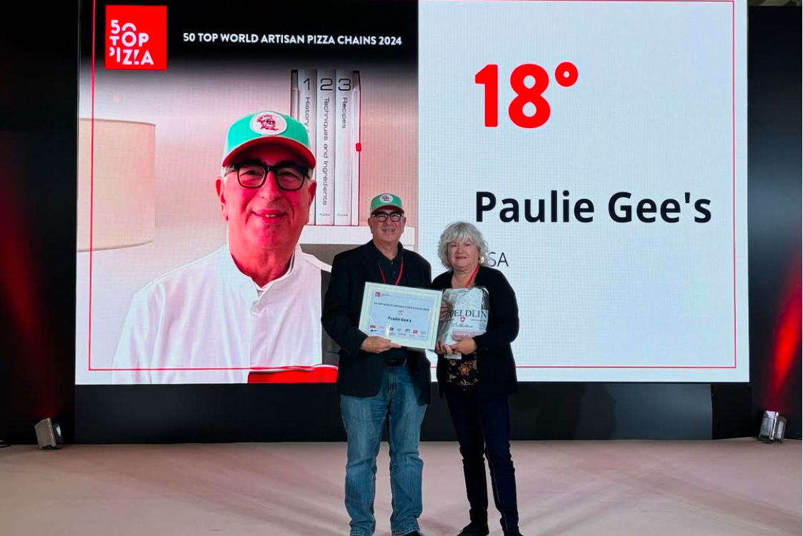 This photo shows Paulie Gee accepting his 50 Top Pizza award with a woman standing next to him. Behind him is a large screen with his face, listing Paulie Gee's as the No. 18 best artisan pizza chain in the world.