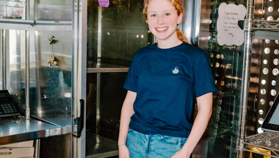 This photo shows a young woman with curly reddish-blonde hair posing in her uniform shirt at Old Scratch Pizza.