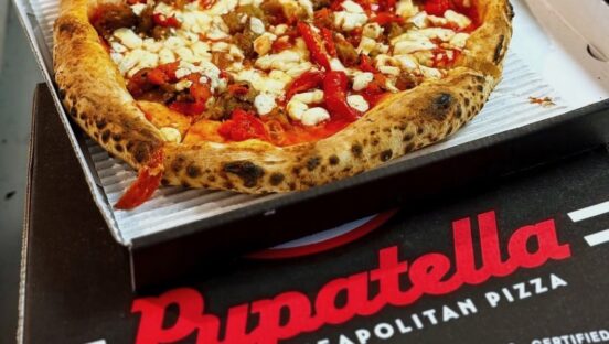 This photo shows a branded Pupatella pizza box containing a pizza featuring cheese, sausage and red peppers.