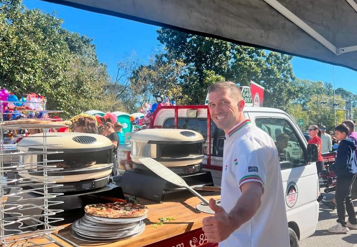 A pizza maker stands at a booth at a pizza festival and smiles toward the camera.