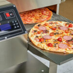 This photo shows a pizza going into a computerized pizza oven, with the high-tech control board next to the conveyor.