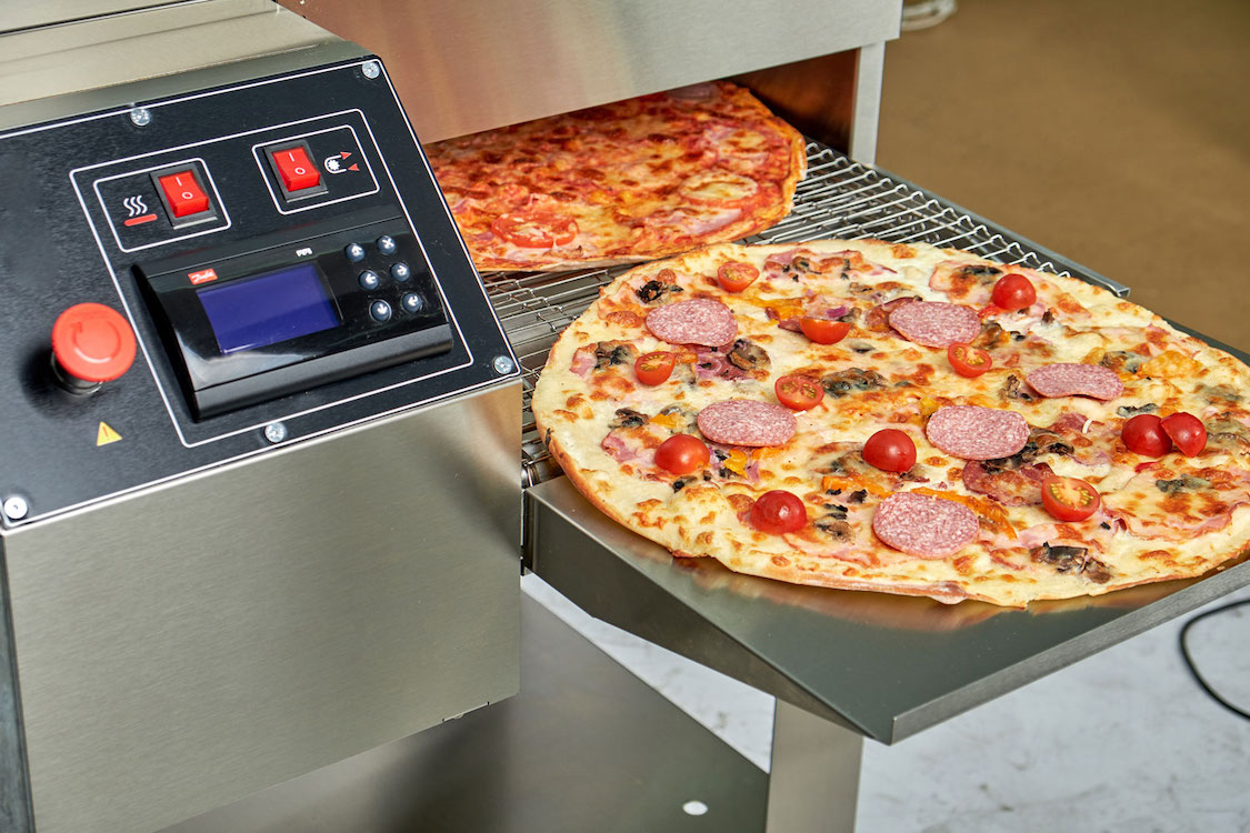 This photo shows a pizza going into a computerized pizza oven, with the high-tech control board next to the conveyor.