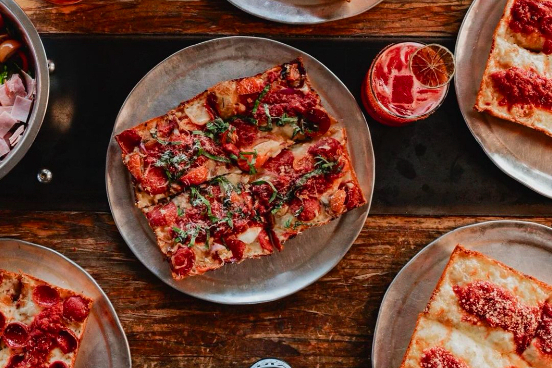 This photo shows a beautiful square-shaped Detroit-style pizza with cheese, sauce, tomato slices and strips of basil.