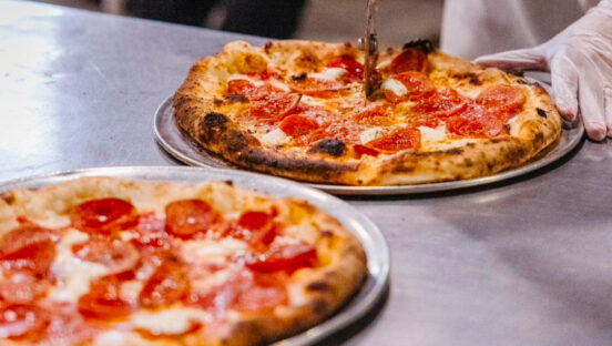 This photo shows a slicer cutting a Neapolitan style pizza on a tray, with another pizza, out of focus, in the foreground.