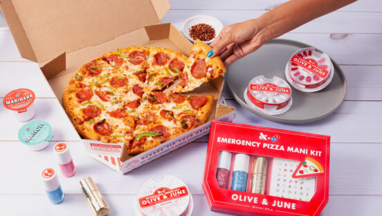 This photo shows a woman's hand, with manicured nails, pulling a slice of pepperoni pizza from a whole Domino's pie, with the Emergency Pizza Mani Kit from Olive & June next to the pizza.