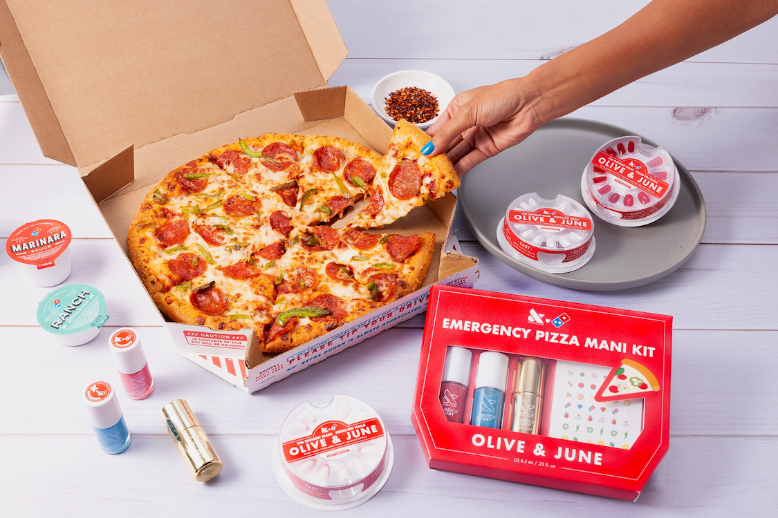 This photo shows a woman's hand, with manicured nails, pulling a slice of pepperoni pizza from a whole Domino's pie, with the Emergency Pizza Mani Kit from Olive & June next to the pizza.