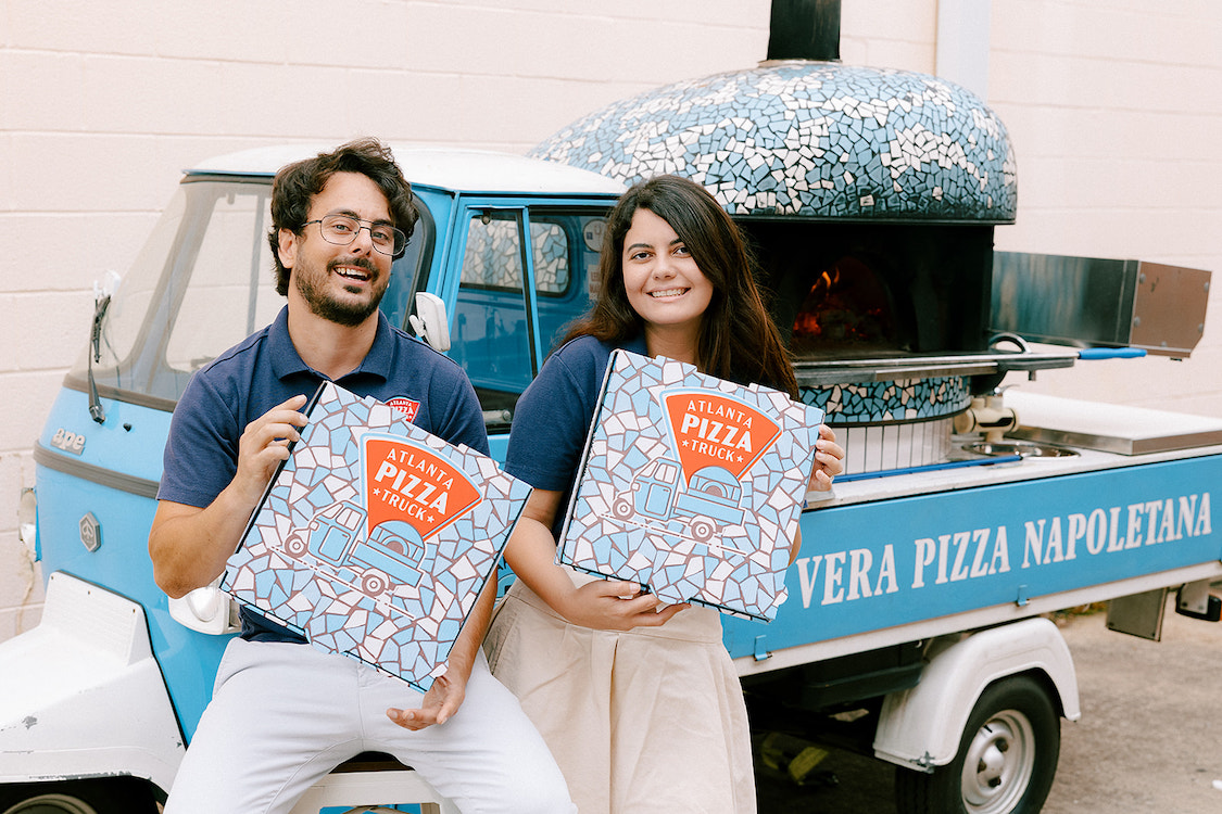 This photo shows Alessio Lacco and Sofia Arango holding up their pizza boxes in front of their blue pizza truck with an oven on the back.