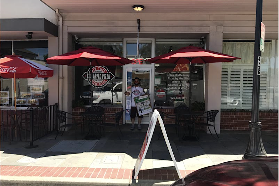 This photo shows a man, seen from a distance, standing in front of the entrance to Big Apple Pizza. He appears to be holding large signs in both hands.