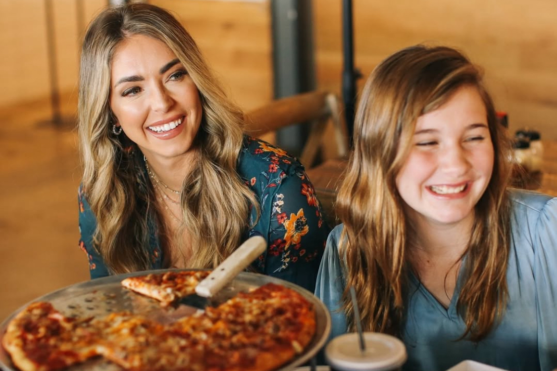This photo shows a woman with long blonde hair and her brown-haired daughter eating a pizza at Crust Pizza Co.