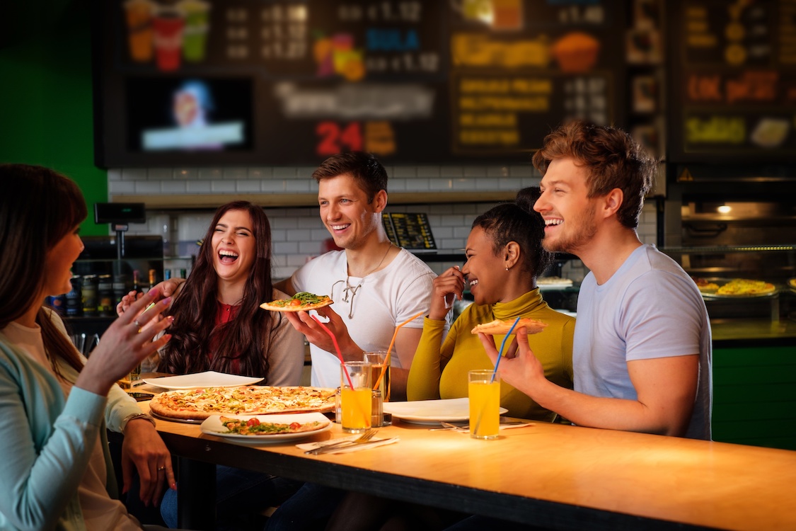 This photo shows a group of cheerful multiracial friends having fun eating pizza in a pizzeria.
