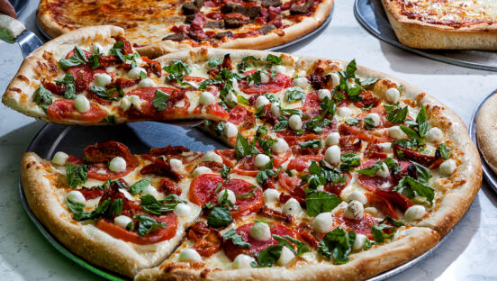 This photo shows a beautiful pizza topped with pepperoni, basil leaves and little balls of mozzarella. Two other pizzas with various toppings can be seen in the background.