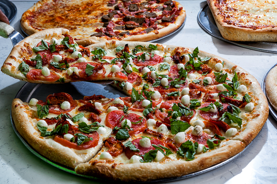 This photo shows a beautiful pizza topped with pepperoni, basil leaves and little balls of mozzarella. Two other pizzas with various toppings can be seen in the background.