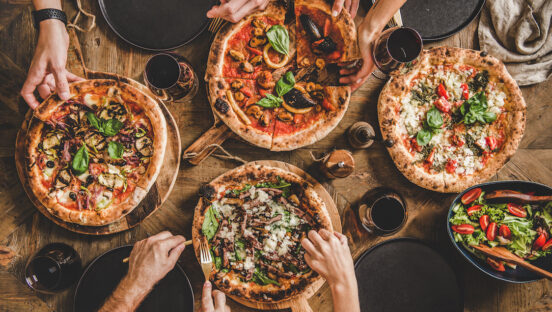 This photo shows the hands of several people pulling out slices from five whole pizzas. There's also a salad on the far right side.