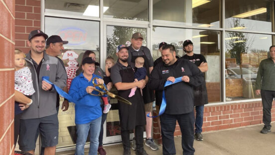 This photo shows Michael LaMarca, wearing a black chef coat, right after he and a team member cut the ribbon at the grand opening of a Master Pizza location. Standing nearby are several other employees, most wearing black shirts, and one of them is holding a baby.