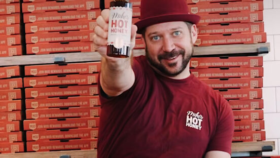 This photo shows Mike Kurtz holding up a bottle of Mike's Hot Honey. He's standing in front of several stacks of MOD Pizza boxes.