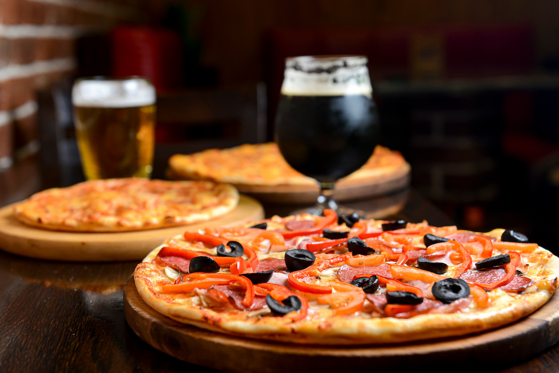 This photo shows a glass of dark beer with a pizza topped with red peppers and black olives, with two more pizzas and a lighter beer in the background.
