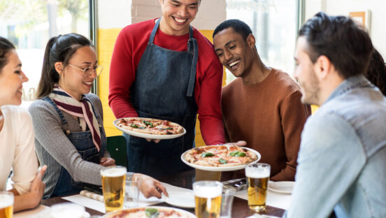 This photo shows a pizzeria server bringing pizzas to a diverse group of customers sitting at a pizzeria table.