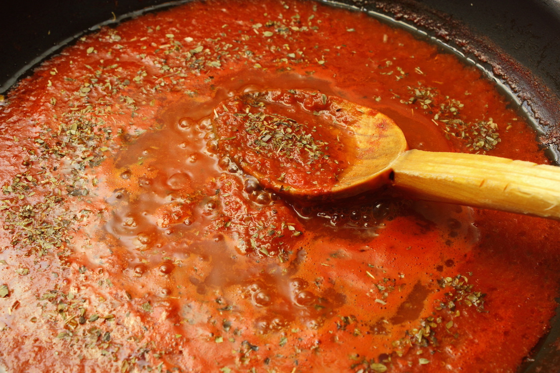 This photo shows a wooden spoon stirring a pot of pizza sauce sprinkled with generous amounts of oregano.