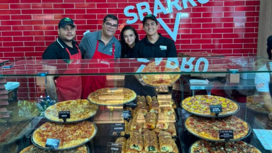 This photo shows a group of Sbarro employees standing and smiling behind a display loaded with whole pizzas.