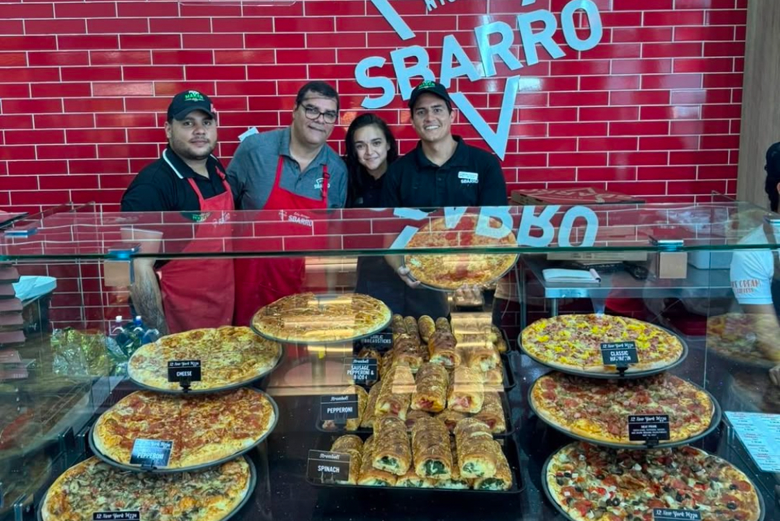 This photo shows a group of Sbarro employees standing and smiling behind a display loaded with whole pizzas.