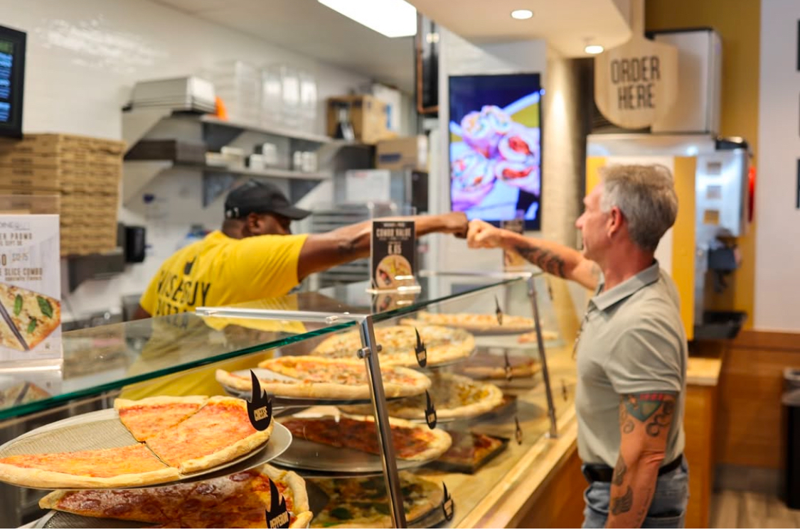 This photo shows a Wiseguy Pizza employee fist-bumping with a customer at the slice counter.